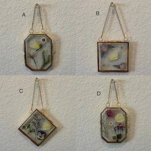 May include: Four decorative pressed flower frames with gold-colored metal edges and hanging chains. Each frame contains dried flowers and petals in various shapes and colors, including yellow, purple, and pink. The frames are displayed against a neutral-colored wall.