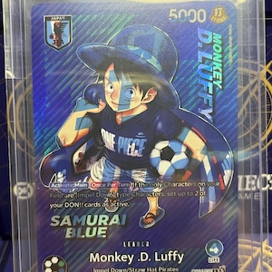May include: A One Piece trading card featuring Monkey D. Luffy in a blue soccer uniform, holding a soccer ball. The card has a blue holographic finish, text including 'SAMURAI BLUE' and 'Monkey D. Luffy', a 5000 power rating, and a 4 life point indicator.