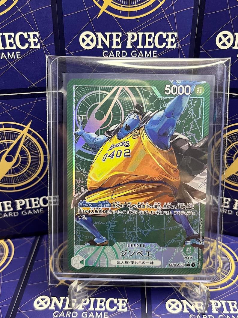 May include: A One Piece card featuring Jinbe in a Lakers jersey. The card has a green background with Japanese text and a 5000 attack power indicator. The card is encased in a protective sleeve.