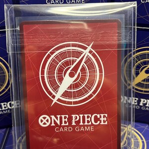 May include: A red One Piece card game card with a white compass design and the text "ONE PIECE CARD GAME." The card is protected in a clear plastic sleeve. Other card game boxes are visible in the background.