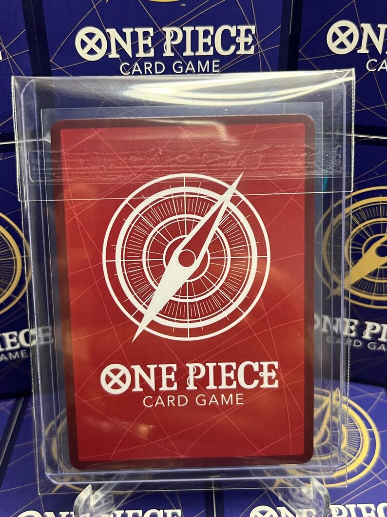 May include: A red One Piece card game card with a white compass design and the text "ONE PIECE CARD GAME." The card is protected in a clear plastic sleeve. Other card game boxes are visible in the background.