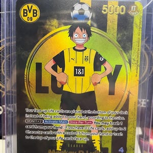 May include: A trading card featuring a cartoon character in a yellow and black soccer jersey with the text "LUCKY". The card has a soccer ball on top and the text "BVB 09" and "1&1". The card also has the text "Monkey.D.Luffy" and "LEADER".