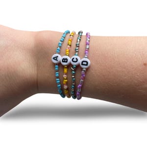 Può includere: Quattro braccialetti di perline nei toni del blu, giallo, verde e viola sono indossati su un polso. Ogni braccialetto ha una perla bianca con una lettera: A, B, C e D. I braccialetti sono fatti di piccole perline colorate.