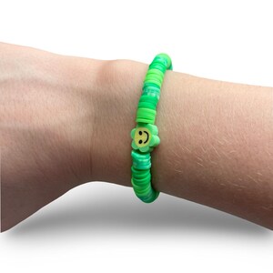 Puede incluir: Una pulsera verde con un dije de flor. La pulsera está hecha de pequeñas cuentas redondas verdes en varios tonos. Un dije de flor verde con una carita sonriente amarilla está centrado en la pulsera. La pulsera se lleva en una muñeca.