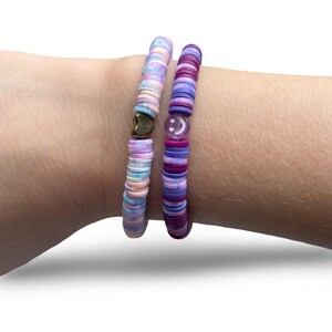 Puede incluir: Dos pulseras de cuentas. Una pulsera presenta cuentas de colores pastel con un dije de corazón dorado. La otra pulsera tiene cuentas moradas y rosas con un dije de carita sonriente. Las pulseras están apiladas en una muñeca.