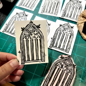May include: Black and white linocut prints of a gothic arched window design. The prints are on off-white paper, with a carved linoleum block visible. The image is on a green cutting mat.