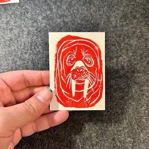May include: A red and white linocut print of a walrus. The walrus has large tusks and a detailed face. Several other prints are visible in the background.
