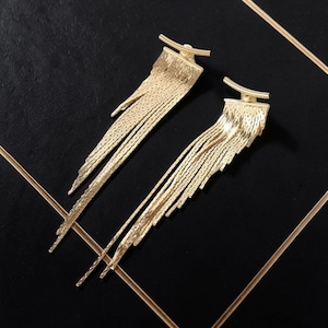 Peut inclure: Une paire de boucles d'oreilles dorées avec un design à franges. Les boucles d'oreilles ont une section supérieure texturée avec une barre incurvée et plusieurs brins de franges pendantes. Les boucles d'oreilles sont présentées sur une surface noire avec des lignes dorées.