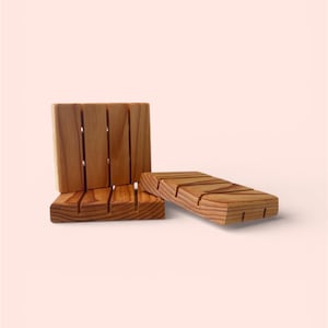 May include: Three wooden soap dishes in a light brown color. Two are rectangular with slotted tops, and one is upright with vertical slots. The soap dishes are made of wood and are designed to hold bars of soap.