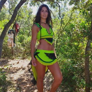 Puede incluir: Un conjunto de dos piezas verde neón con detalles de encaje negro. La parte superior tiene un diseño de cuello halter, y la falda es corta con un dobladillo con volantes. El conjunto se usa al aire libre, rodeado de árboles y follaje.