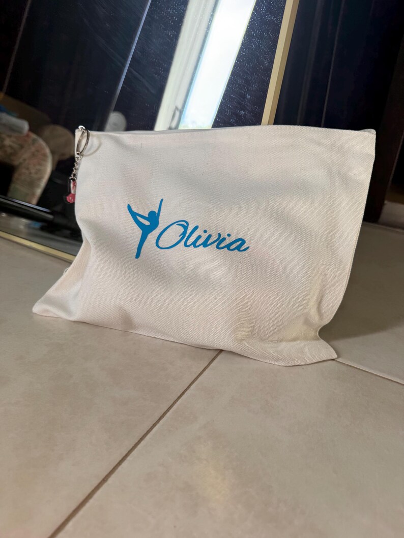 Peut inclure: Trousse de maquillage en toile blanche avec fermeture &eacute;clair. Le sac pr&eacute;sente un motif bleu d'une gymnaste et le nom "Olivia" en cursive. Un petit charme est accroch&eacute; &agrave; la tirette.