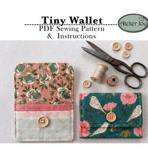 May include: Two fabric wallets, one with a pink floral pattern and one with a teal bird and flower print, each featuring a wooden button closure. Includes sewing supplies: metal scissors, wooden spools, and buttons. Text reads: Tiny Wallet PDF Sewing Pattern & Instructions, Atelier Joy.