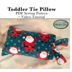 May include: A green and red tie pillow with a Santa Claus pattern. The pillow is tied with a green and red fabric tie. The pillow is on a beige surface. "Toddler Tie Pillow" and "PDF Sewing Pattern + Video Tutorial" are written in black text above the pillow. "Atelier Joy" is written in a circle with a green background and white text.