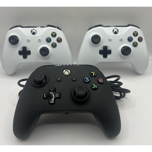 Puede incluir: Se muestran tres mandos de Xbox sobre un fondo blanco. Dos son blancos y uno negro. Cada mando tiene un panel direccional, dos palancas analógicas y botones de colores. El mando negro tiene un cable.