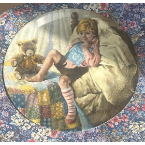 Könnte beinhalten: Dekorative Platte mit einem Kind im Bett, das ein Buch mit dem Titel "Dear Stories" hält. Ein Teddybär sitzt daneben auf einer Patchwork-Decke. Die Platte hat einen floralen Rand und einen Vintage-Stil.