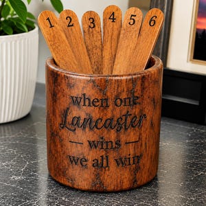 May include: A cylindrical brown wood-grain container holding six wooden sticks numbered one through six. The container features the engraved text "when one Lancaster wins we all win" in a dark font on its front surface.