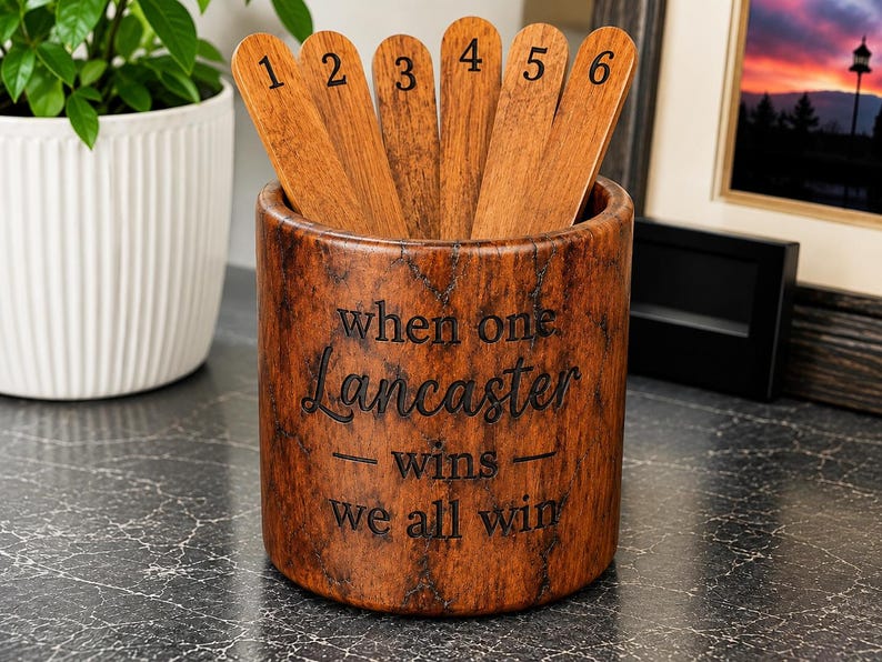 May include: A cylindrical brown wood-grain container holding six wooden sticks numbered one through six. The container features the engraved text "when one Lancaster wins we all win" in a dark font on its front surface.