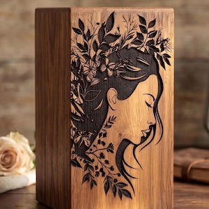 A wooden urn box featuring a dark laser-engraved profile of a woman with floral and leafy hair decorations.