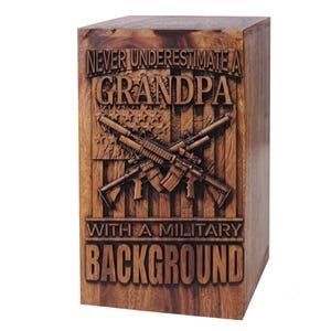 A wooden urn box carved with an American flag, crossed rifles, and the text NEVER UNDERESTIMATE A GRANDPA WITH A MILITARY BACKGROUND.