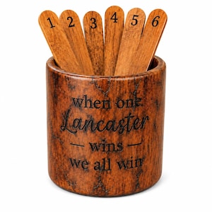 May include: A cylindrical brown wood-grain container holding six wooden sticks numbered one through six. The container features the engraved text: when one Lancaster wins we all win. The wood has a dark, mottled, and textured finish throughout.