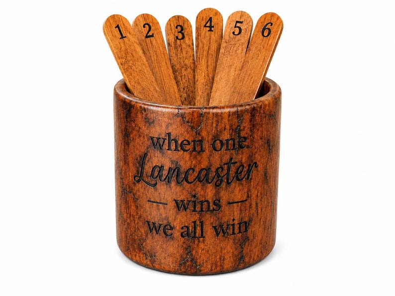 May include: A cylindrical brown wood-grain container holding six wooden sticks numbered one through six. The container features the engraved text: when one Lancaster wins we all win. The wood has a dark, mottled, and textured finish throughout.