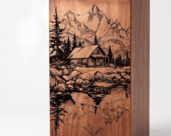 May include: A rectangular wooden box featuring a dark, laser-engraved illustration of a cabin nestled among pine trees and rocky terrain, set against a backdrop of jagged mountains with a clear reflection of the scene visible in the water below.