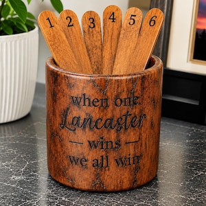 May include: A cylindrical brown wood-grain container holding six wooden sticks numbered one through six. The container features the engraved text "when one Lancaster wins we all win" in a dark font on its front surface.
