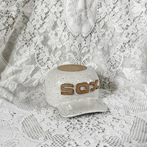 May include: A ceramic baseball cap sculpture with a speckled white glaze and a brown clay finish on the top opening. The front of the cap features the letters SOX in raised brown clay lettering, and the piece includes a curved brim at the base.