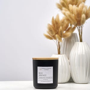 May include: A black candle with a wooden lid and a label that reads "Blue Serenity" sits on a white surface. Behind it are three white vases with dried, fluffy, tan-colored flowers. The candle is from Inspire & Company Candles.