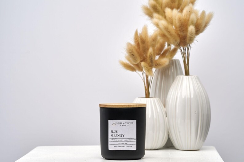 May include: A black candle with a wooden lid and a label that reads "Blue Serenity" sits on a white surface. Behind it are three white vases with dried, fluffy, tan-colored flowers. The candle is from Inspire & Company Candles.