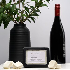 May include: A black vase with green leafy branches, a black framed tin of "Blue Serenity" candles, a bottle of red wine, and several white wax cubes. The items are arranged on a white surface.