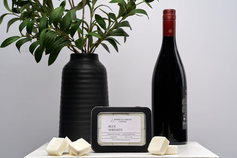 May include: A black vase with green leafy branches, a black framed tin of "Blue Serenity" candles, a bottle of red wine, and several white wax cubes. The items are arranged on a white surface.