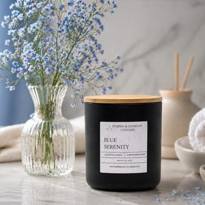 May include: A black candle with a wooden lid labeled "Blue Serenity" sits on a marble surface. A clear glass vase with blue flowers, a rolled white towel, and a bowl of blue crystals are also present, creating a spa-like ambiance.