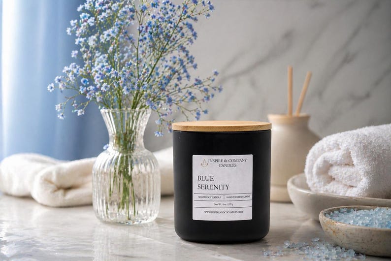 May include: A black candle with a wooden lid labeled "Blue Serenity" sits on a marble surface. A clear glass vase with blue flowers, a rolled white towel, and a bowl of blue crystals are also present, creating a spa-like ambiance.