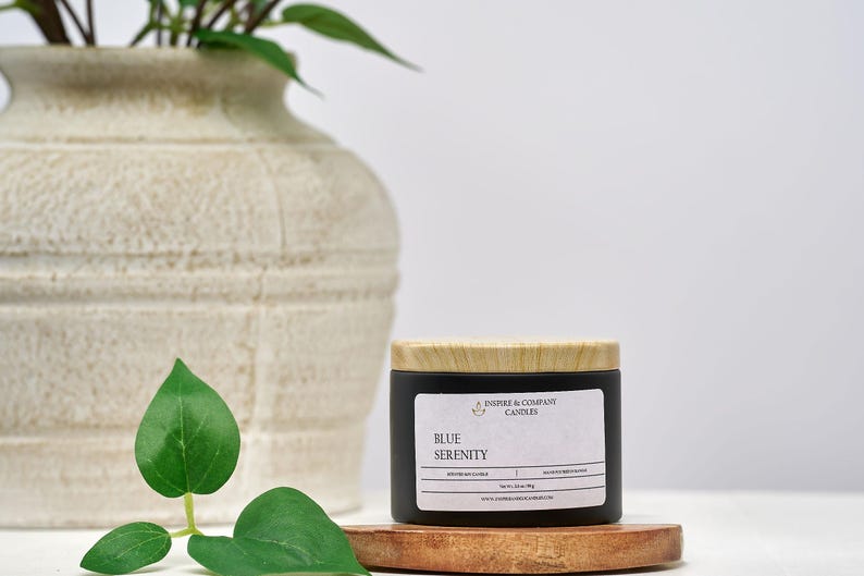 May include: A black candle jar with a wooden lid, labeled "Blue Serenity." The candle sits on a small wooden tray, with a large off-white vase and green leaves in the background. The candle is from Inspire & Company Candles.