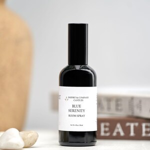 May include: A black room spray bottle with a white label that reads "Blue Serenity Room Spray." The bottle is next to white stones and a tan vase. The background includes books with the words "Create" and "Inspiration."