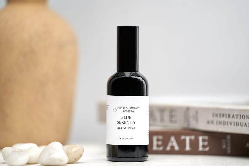 May include: A black room spray bottle with a white label that reads "Blue Serenity Room Spray." The bottle is next to white stones and a tan vase. The background includes books with the words "Create" and "Inspiration."