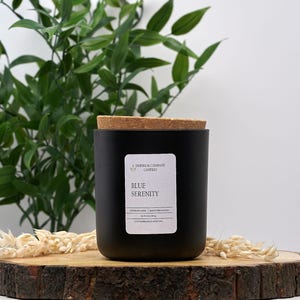May include: A black candle jar with a cork lid, labeled "Blue Serenity." The jar sits on a wooden slice, with green foliage and dried flowers in the background. The candle is a home decor item.