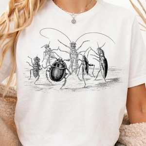Puede incluir: Una camiseta blanca de manga corta con una detallada ilustración en tinta negra de cinco insectos diferentes dispuestos en círculo. El gráfico muestra varios escarabajos y cucarachas con trazos finos, evocando un estilo de grabado antiguo.