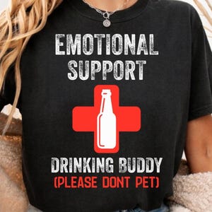 Op de afbeelding: Zwart t-shirt met de tekst "EMOTIONAL SUPPORT" boven een rood kruis met een witte fles. Daaronder staat "DRINKING BUDDY (PLEASE DONT PET)".