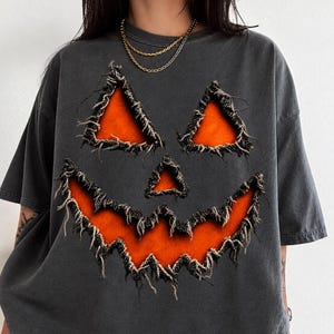 Może przedstawiać: Ciemnoszary t-shirt z krótkim rękawem z dużym, postarzanym nadrukiem twarzy dyni. Oczy, nos i poszarpane usta są w kolorze jaskrawego pomarańczu, z postrzępionymi, surowymi krawędziami materiału wokół ciemnych konturów wzoru.