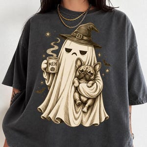 May include: A charcoal gray short-sleeve t-shirt featuring a graphic of a ghost wearing a witch hat, holding a mug with the text MEH and a pumpkin icon, while carrying a French bulldog. The design includes small stars and bats in a beige tone.