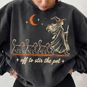 Puede incluir: Una sudadera de cuello redondo gris carbón con un estampado gráfico de una bruja sosteniendo una escoba, seguida de tres gatos. Arriba hay una luna creciente naranja y estrellas. El texto de abajo dice, off to stir the pot.