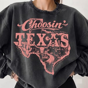 Può includere: Una felpa girocollo grigio antracite con una grafica rosa sbiadita dello stato del Texas. Il design include il testo "Choosin' TEXAS" con illustrazioni di un teschio di longhorn, un cactus, una stella e una strada tortuosa all'interno del Texas.
