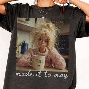 K&ouml;nnte beinhalten: Dunkelgraues T-Shirt mit einem Puppenbild, das einen rosa Pullover tr&auml;gt und eine Tasse mit der Aufschrift "TEACH" h&auml;lt. Der Text "made it to may" steht darunter. Ein l&auml;ssiger, bequemer Stil.