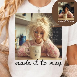 K&ouml;nnte beinhalten: Wei&szlig;es T-Shirt mit einer Illustration einer Puppe mit zerzaustem blondem Haar, einem rosa Pullover und einer Tasse mit der Aufschrift "TEACH". Der Text "made it to may" ist darunter gedruckt. Ein kleineres Bild der Puppe befindet sich in der Ecke.