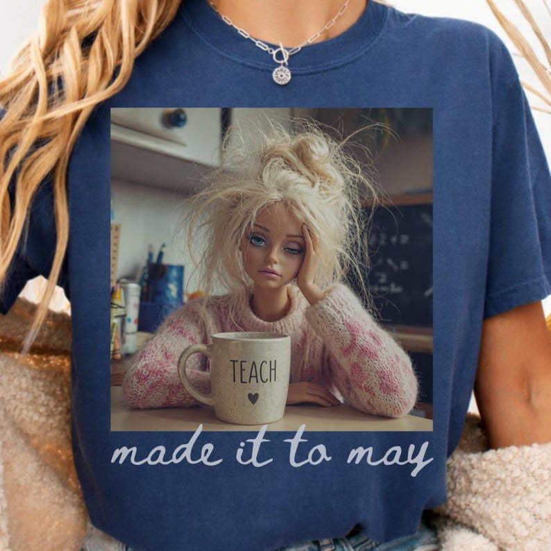 K&ouml;nnte beinhalten: Marineblaues T-Shirt mit einem Aufdruck einer Puppe mit zerzaustem blondem Haar, die ihren Kopf auf ihre Hand st&uuml;tzt. Die Puppe h&auml;lt eine Tasse mit der Aufschrift "TEACH" und einem Herz. Der Text "made it to may" steht darunter.