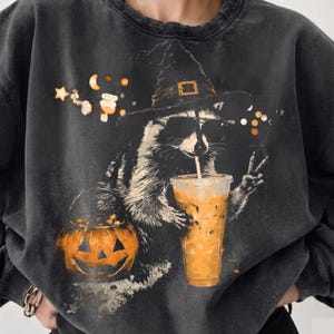 May include: A charcoal gray mineral-washed sweatshirt featuring a graphic of a raccoon wearing a witch hat and sunglasses, holding an iced coffee and making a peace sign. An orange jack-o'-lantern bucket sits nearby with stars and moons.