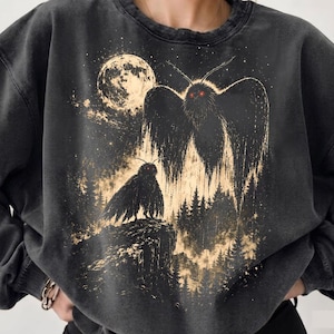 Könnte beinhalten: Ein anthrazitfarbenes, mineralgewaschenes Sweatshirt mit Rundhalsausschnitt und einem Grafikdruck von zwei dunklen, geflügelten Kreaturen mit leuchtend roten Augen. Das Motiv zeigt einen Vollmond, einen dichten Kiefernwald und eine Felsklippe.