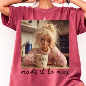 K&ouml;nnte beinhalten: Burgunderfarbenes T-Shirt mit einem aufgedruckten Bild einer Puppe mit zerzaustem blondem Haar, die ihren Kopf auf ihre Hand st&uuml;tzt. Sie tr&auml;gt einen rosa Pullover und h&auml;lt eine Tasse mit der Aufschrift "TEACH". Der Text "made it to may" ist darunter gedruckt.